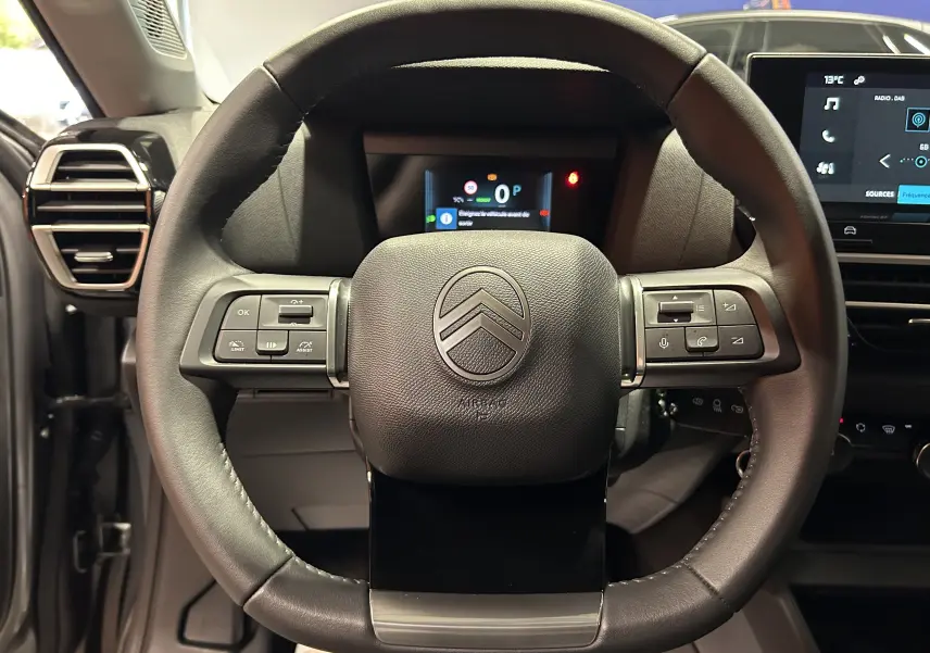 Vue frontale du volant noir du Citroën E-C4 électrique 2025 avec commandes intégrées et tableau de bord numérique en arrière-plan.