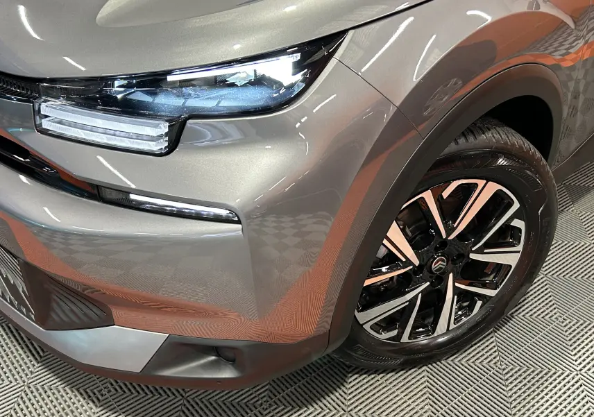 Gros plan sur l'avant droit gris foncé du Citroën E-C4 2025, mettant en valeur le phare LED et la jante alliage bicolore.