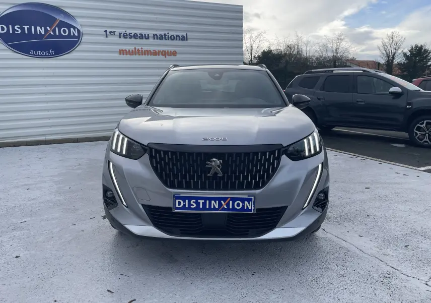 Vue frontale d'un Peugeot 2008 gris Artense avec calandre noire et feux LED distinctifs allumés, sur parking extérieur.