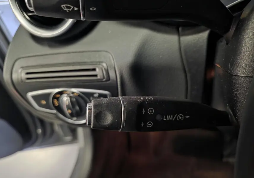 Gros plan sur la commande de régulateur de vitesse à gauche du volant dans l’habitacle noir d’une Mercedes Classe C gris métallisé.