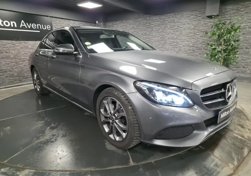 Mercedes Classe C gris métallisé vue 3/4 avant droit avec phares LED allumés en intérieur showroom.