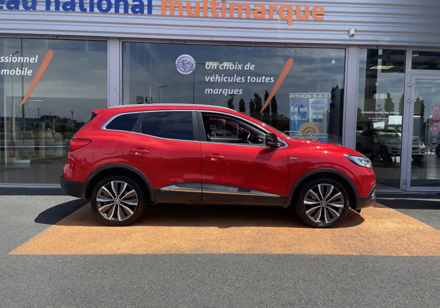 Profil droit d'un Renault Kadjar rouge flamme 2018 avec jantes alliage bi-ton et vitres teintées, devant une vitrine commerciale.