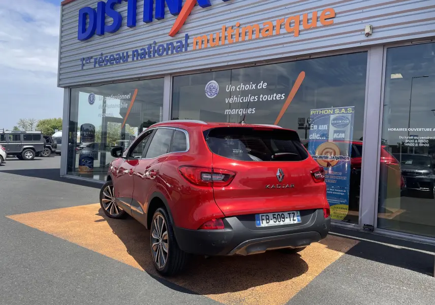 Vue 3/4 arrière droite d’un Renault Kadjar rouge flamme garé devant un showroom multimarque avec jantes alliage bicolores.