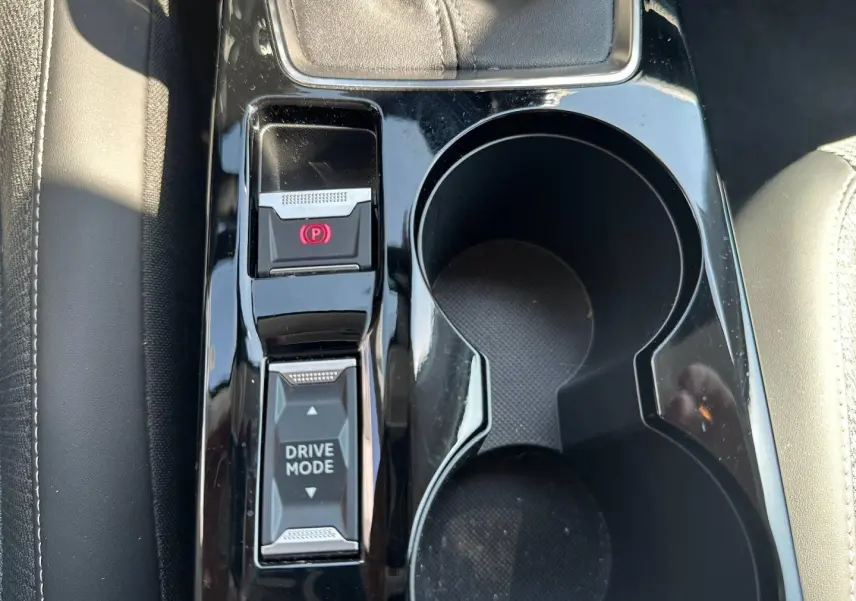 Gros plan sur la console centrale noire brillante avec bouton frein à main électrique et sélecteur Drive Mode, sièges gris visibles.