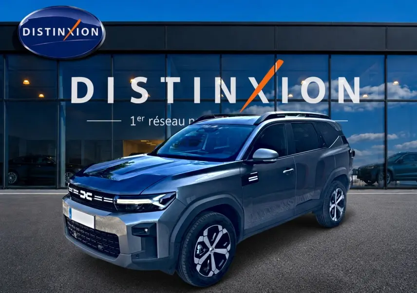 SUV Dacia Bigster gris schiste en 3/4 avant droit, avec jantes alu et barres de toit visibles devant un showroom Distinxion.