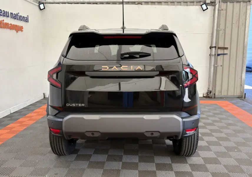 Vue arrière du Dacia Duster noir 2025 avec logo doré et feux arrière en forme de C, stationné en intérieur.