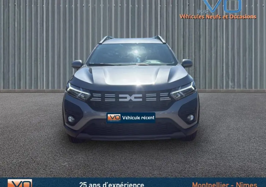 Vue frontale d'un Dacia Jogger gris 2025 avec barres de toit et feux LED allumés sur un parking.