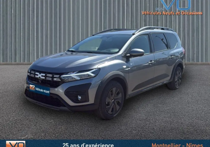Vue 3/4 avant droite d'un Dacia Jogger gris 2025 avec barres de toit et jantes noires, stationné devant un mur métallique.