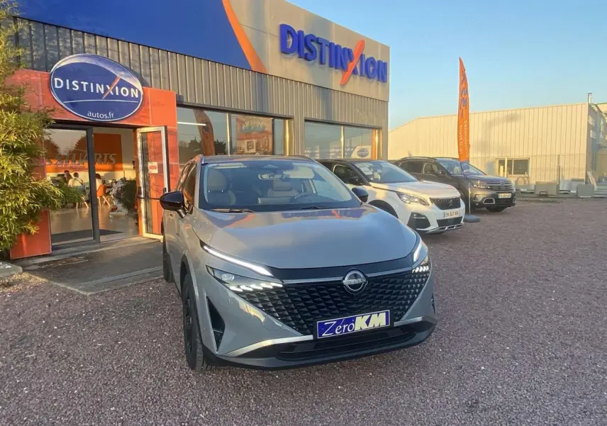 Vue 3/4 avant d'un Nissan Qashqai gris ceramic 2025 garé devant un concessionnaire avec plaque "Zéro KM".