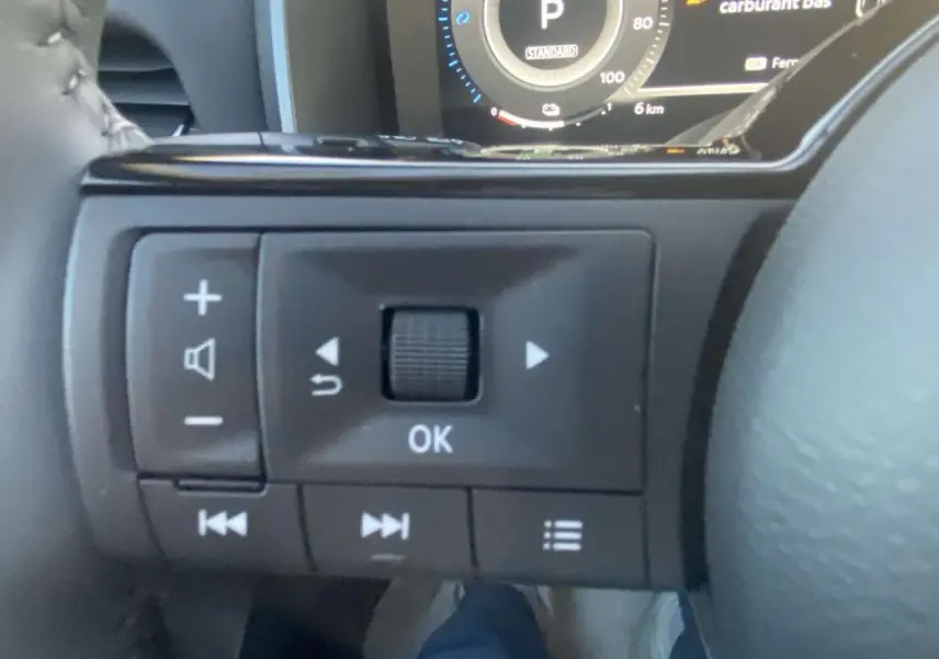 Gros plan sur les commandes au volant côté gauche du Nissan Qashqai gris Ceramic, tableau de bord numérique en arrière-plan.