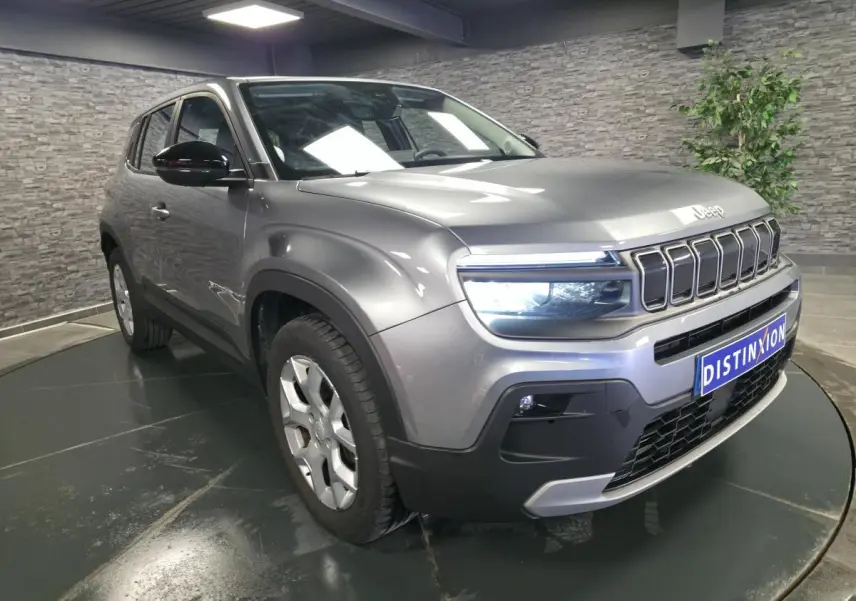 Jeep Avenger gris métallisé en 3/4 avant droit, avec calandre distinctive et phares LED allumés, en intérieur showroom.