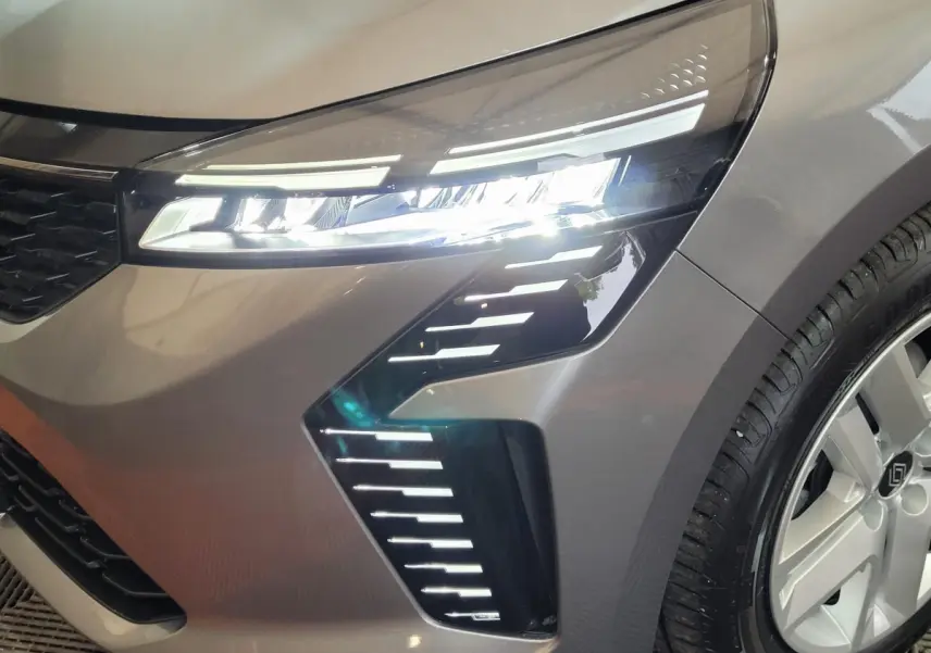 Gros plan sur le phare avant droit allumé d'une Renault Clio gris foncé, avec jante et partie de la calandre visibles.