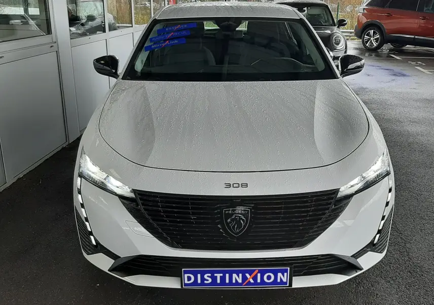 Vue avant d'une Peugeot 308 blanche Banquise avec phares allumés et plaque Distinxion visible.