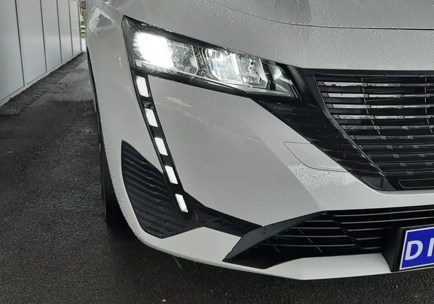 Gros plan sur l'avant gauche blanc banquise d'une Peugeot 308 2022 avec phares LED allumés et calandre noire.