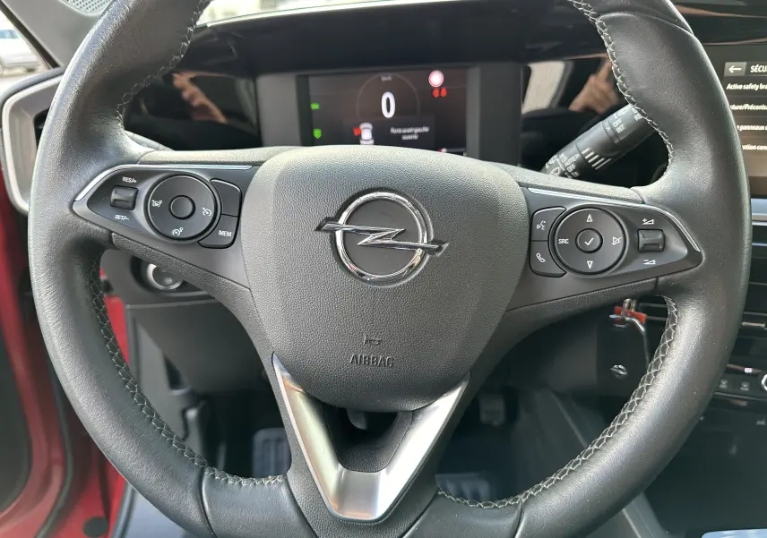 Gros plan sur le volant en cuir noir de l'Opel Mokka rouge Kardinal, avec commandes intégrées et tableau de bord numérique visible.