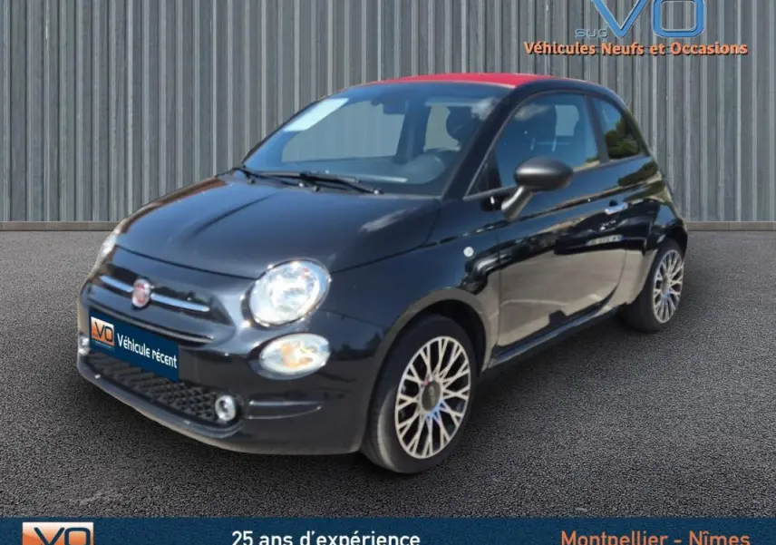 Vue 3/4 avant droite d'une FIAT 500C noire 2024 avec toit rouge et jantes alliage, stationnée sur sol bitumé.