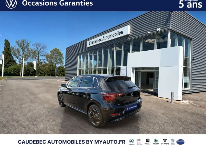 Vue 3/4 arrière droite d'une Volkswagen Polo noire intense nacrée, stationnée devant un bâtiment commercial moderne.