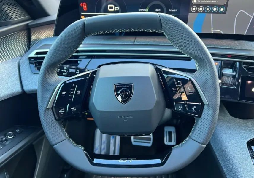 Vue rapprochée du volant cuir perforé gris du Peugeot 3008 Hybrid GT avec logo lion et inscription GT, tableau de bord digital en arrière-plan.