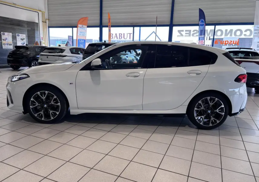BMW Série 1 120d M Sport blanc vue de profil côté gauche en intérieur, avec jantes alliage noires et vitres teintées.