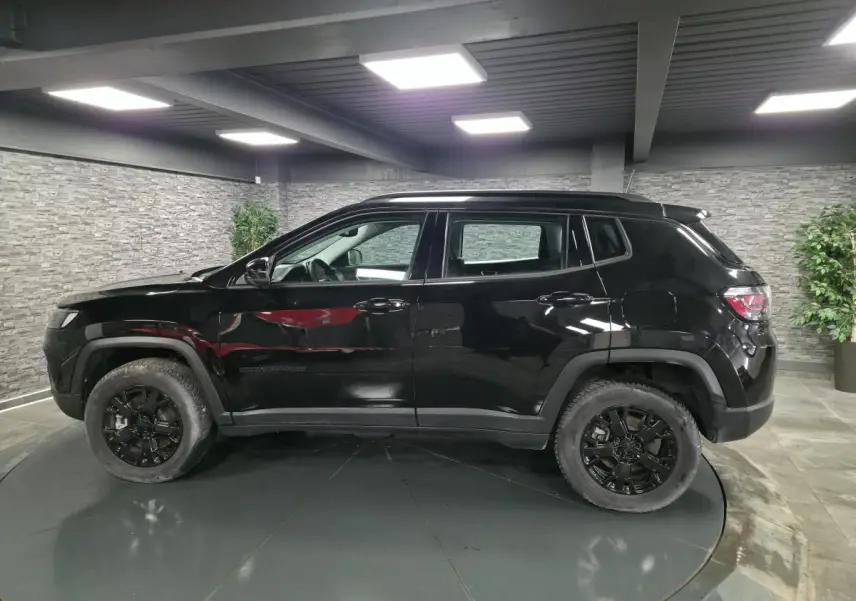 Profil côté gauche du Jeep Compass noir 2024 avec jantes noires et garde au sol surélevée en intérieur showroom.