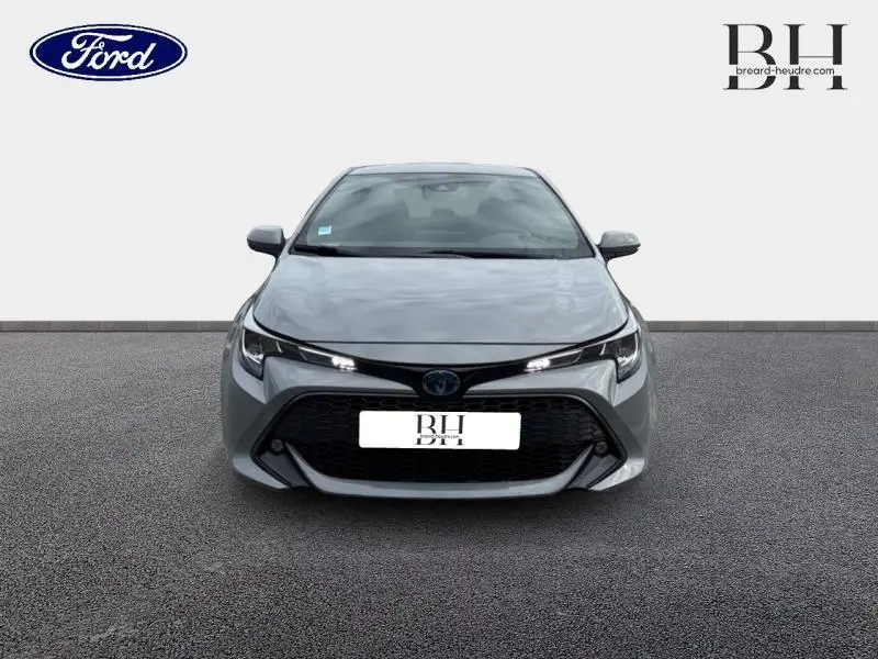 Vue avant d'une Toyota Corolla 122h Dynamic Business 2020 en gris Manhattan métallisé avec calandre noire et phares LED allumés.