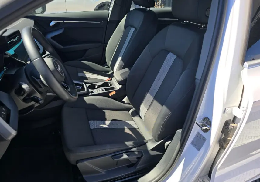 Vue intérieure côté conducteur de l'Audi A3 Sportback 2024 en Blanc Arkona, avec sièges tissu noir et console centrale visible.