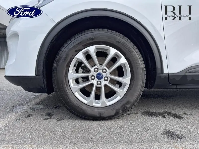 Gros plan sur la roue avant droite d’un Ford Kuga blanc Glacier, mettant en valeur la jante alliage et le logo Ford central.