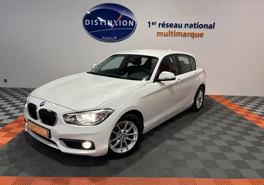BMW Série 1 116d Business blanche vue en 3/4 avant droit, phares allumés, sur sol damier gris et orange.