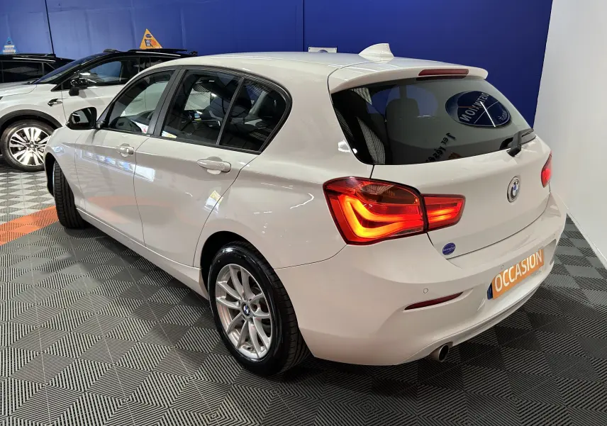 BMW Série 1 116d Business blanche vue 3/4 arrière droit, feux arrière allumés, en très bon état showroom.