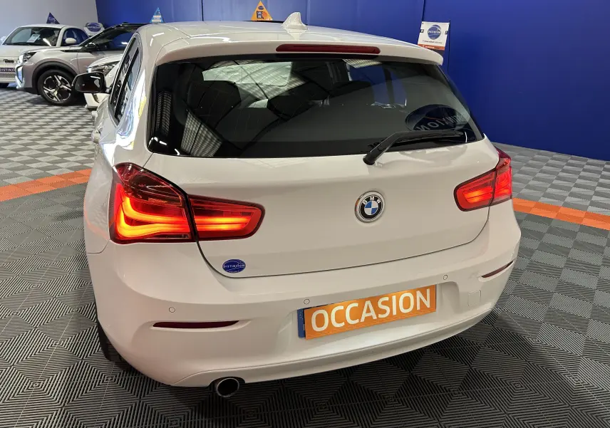 Vue arrière droite d'une BMW Série 1 116d Business blanche de 2015 avec feux allumés dans un showroom.