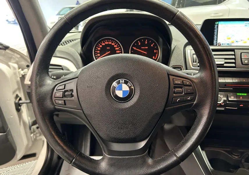 Vue rapprochée du volant noir de la BMW Série 1 116d avec tableau de bord et écran de navigation visibles.