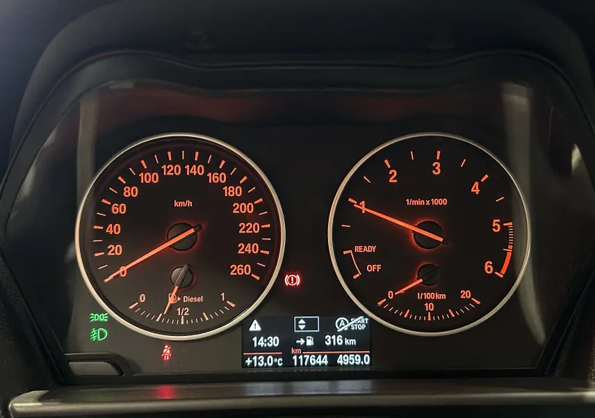 Tableau de bord de BMW Série 1 116d blanc, affichant compteurs vitesse et régime moteur éclairés en rouge.