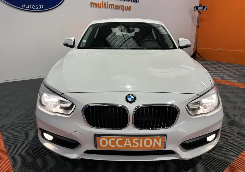 Vue avant d'une BMW Série 1 blanche 116d Business 5 portes avec phares et antibrouillards allumés en showroom.