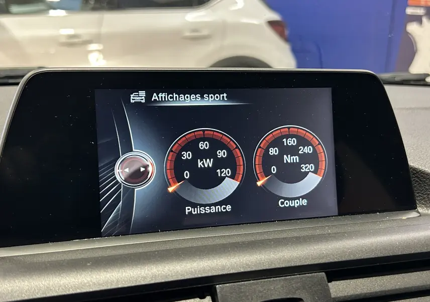 Vue intérieure sur l'écran central affichant les données sportives de puissance et couple dans une BMW Série 1 blanche visible en arrière-plan.