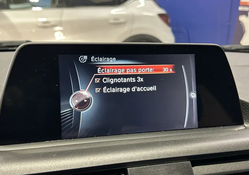 Écran central de la BMW Série 1 116d blanc montrant le menu éclairage avec options clignotants et accueil activées.