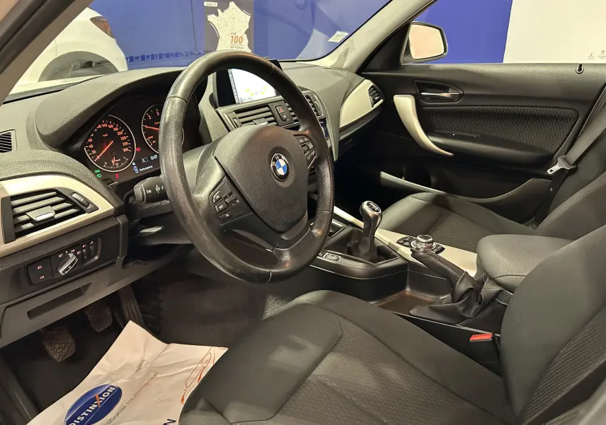 Vue intérieure côté conducteur de la BMW Série 1 116d Business 2015 avec volant cuir, tableau de bord et levier de vitesse manuel.