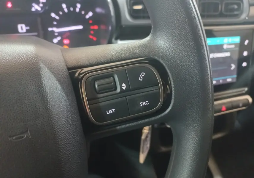Gros plan sur les commandes gauche du volant noir d'une Citroën C3 2024, avec tableau de bord et écran tactile en arrière-plan.