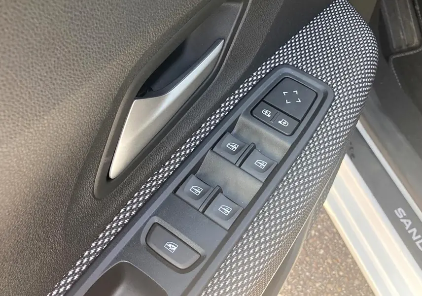 Gros plan sur la commande des vitres électriques côté gauche avec tissu gris à motifs et poignée argentée, portière blanche visible.