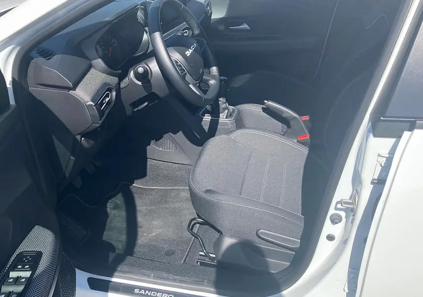 Vue intérieure côté conducteur d'une Dacia Sandero blanche 2025, volant multifonctions et sièges tissu gris.