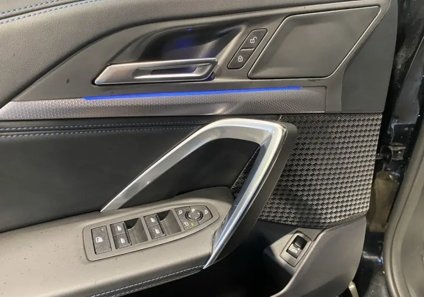 Vue rapprochée de la porte avant gauche noire du BMW X1 2025 avec commandes électriques et éclairage d'ambiance bleu.