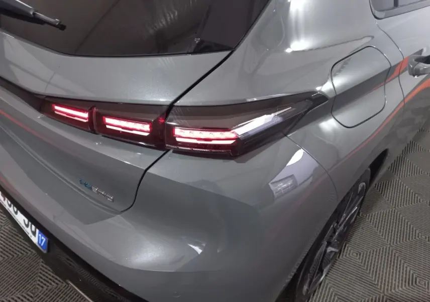 Vue 3/4 arrière droite d'une Peugeot 308 PHEV 180 gris foncé avec feux arrière LED allumés et prise de recharge visible.