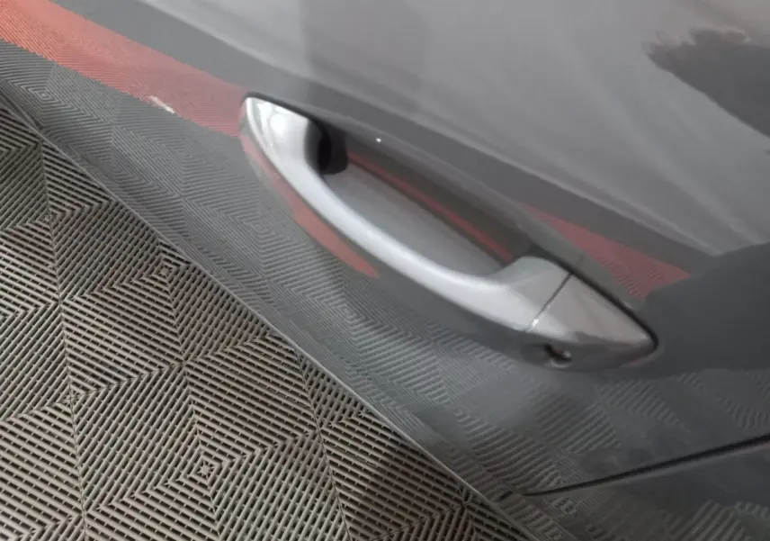 Poignée de porte côté gauche gris foncé de la Peugeot 308 PHEV 180 E-EAT8 Allure 2025, reflet du sol texturé visible.