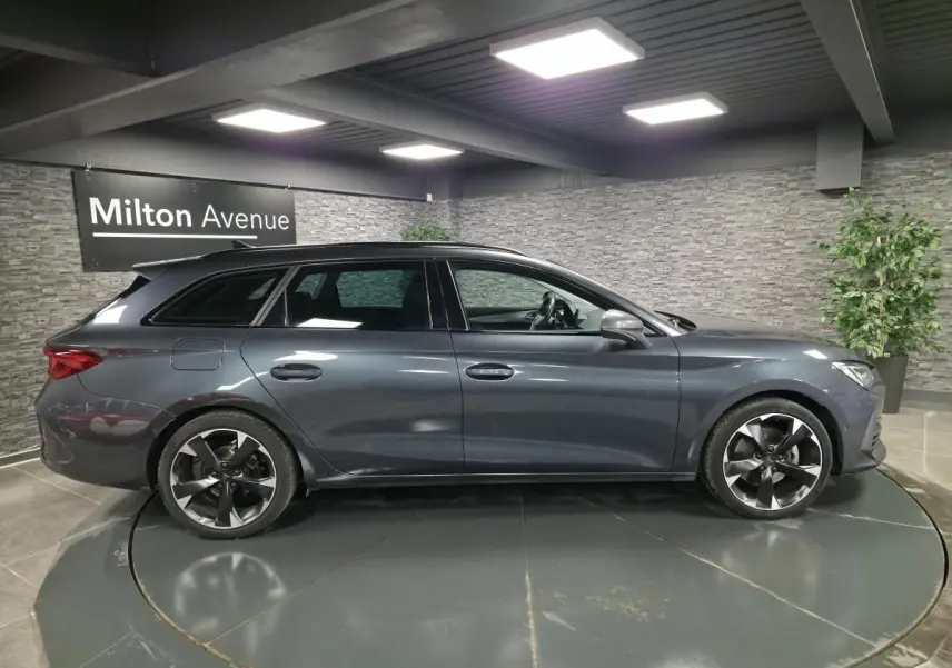 Vue de profil côté gauche d'une CUPRA Leon Sportstourer gris magnétique avec jantes alliage 18 pouces dans un showroom.