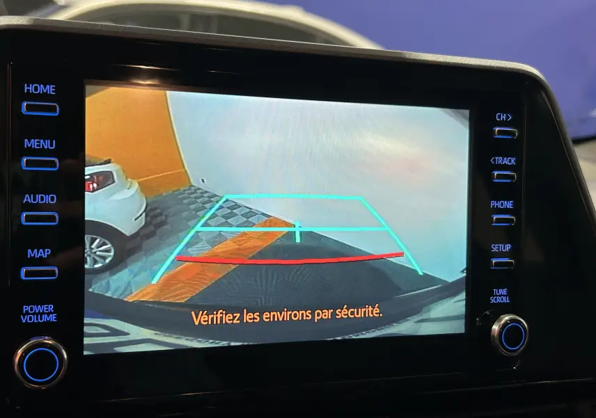 Écran de caméra de recul montrant le sol et un mur orange, avec une voiture blanche garée à gauche.