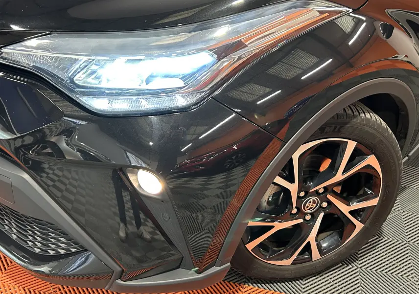 Gros plan sur l'avant droit noir métallisé du Toyota C-HR Hybride 122h avec phare LED allumé et jante bi-ton.