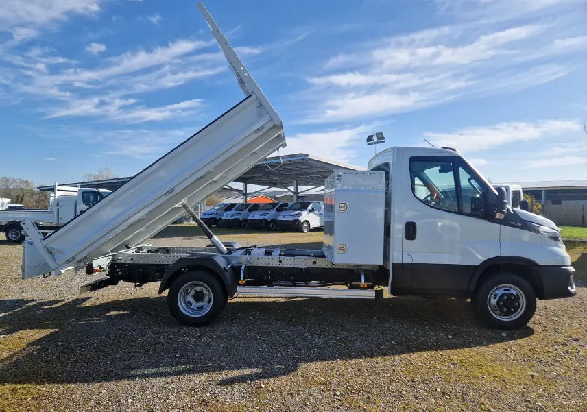 Vue latérale droite d’un Iveco Daily blanc avec benne relevée et coffre latéral acier, sur un terrain extérieur ensoleillé.
