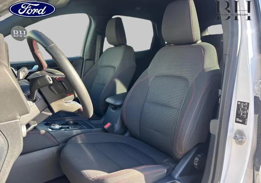 Vue intérieure côté conducteur du Ford Kuga blanc, sièges sport noirs avec surpiqûres rouges et volant cuir multifonction.