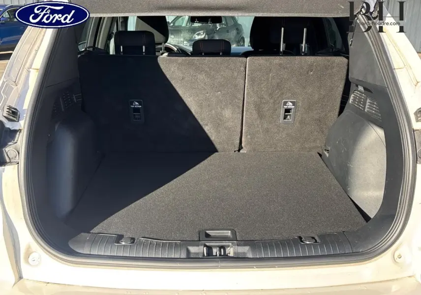 Vue arrière du coffre ouvert d'un Ford Kuga blanc Premium Platinum, montrant la banquette arrière et le tapis de coffre noir.