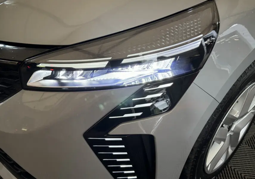 Gros plan sur le phare avant droit allumé d'une Renault Clio gris clair 2025 avec jante et détails LED distinctifs.