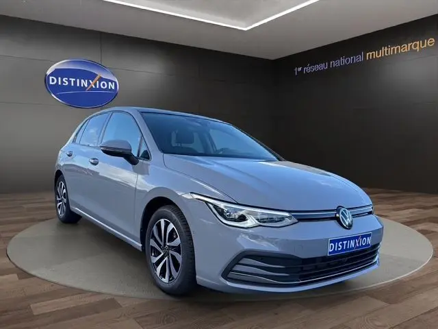 Volkswagen Golf VIII gris lunaire vue 3/4 avant droit dans showroom avec logo Distinxion en arrière-plan.